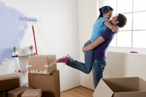 A young couple in casual clothing, with the woman wearing a blue top and jeans and the man wearing a purple t-shirt and jeans, are seen inside a well-lit, modern room near a large window. The man is holding the woman off the ground in an embrace, both smiling and appearing cheerful. The room has a light-colored wall, and there are several cardboard boxes of various sizes, some sealed with packing tape, stacked on the floor. One of the boxes is open, revealing packing materials inside. A rolled-up sheet of bubble wrap is mounted on the wall, along with a red-handled tool or pole leaning against the wall. The scene suggests a move or relocation, with the couple surrounded by packing supplies, in a setting typical of a house or apartment preparing for a household move, possibly in the Haringey area, associated with Haringey Man with Van's removals services.