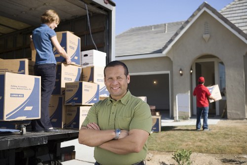 A man with light skin, short dark hair, and a broad smile is standing outdoors in front of a residential property on a clear day, with arms crossed and wearing a green polo shirt. Behind him, two people are involved in a house removal process; one is a young boy with light hair in a blue shirt, stacking or handling moving boxes inside the open back of a large removals van, which is parked on the driveway of a modern, detached house with a light-grey exterior and a sloped roof. The boy is placing or adjusting cardboard boxes, some of which display blue and white branding typical of moving supplies. In the background, another individual dressed in red is carrying a box towards the house entrance. The scene depicts a residential street in Haringey, focused on the logistics of house removals, with the environment clean and well-lit, emphasizing the professional local services provided by Haringey Man with Van.