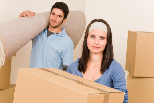 A young man with short dark hair and wearing a light blue shirt is seen in the background, carrying a large, beige fabric-covered sofa over his shoulder inside a house during a move. In the foreground, a young woman with long dark hair, a light grey headband, and wearing a blue top, is holding a medium-sized cardboard box, looking directly at the camera with a neutral expression. She is surrounded by additional moving boxes, some stacked on top of each other, which are made of plain brown cardboard and vary in size. The environment appears to be an interior space with neutral-colored walls, and natural lighting illuminates the scene, suggesting daytime. The setting likely indicates a house or flat being prepared for relocation, with visible elements supporting the context of house removals in Haringey or nearby areas, as referenced on the company's website, haringeymanwithvan.co.uk, specializing in professional removals services.