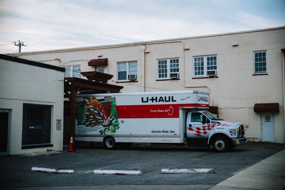 A white U-Haul moving truck with red branding is parked adjacent to a beige, two-storey building with multiple windows, some fitted with air conditioning units. The truck's cargo area displays illustr