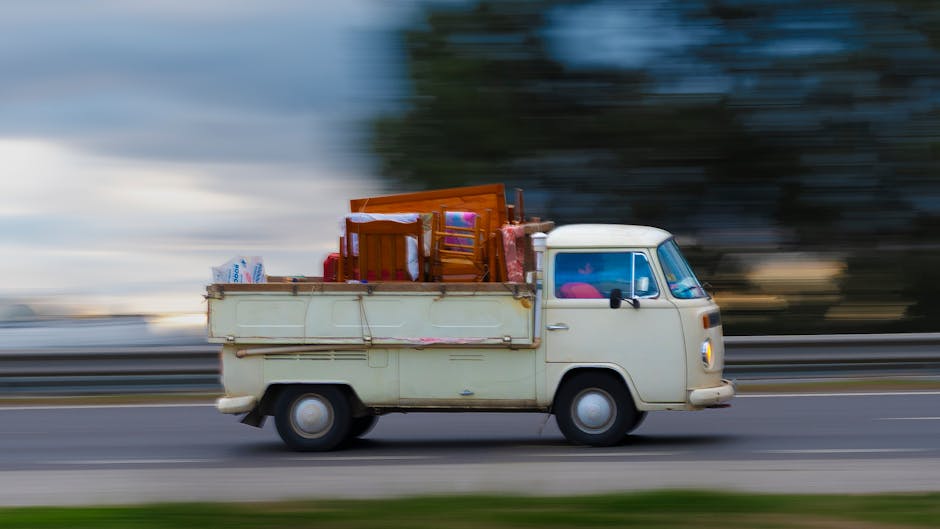 A vintage white delivery van in motion on a roadway, loaded with assorted household furniture and belongings, including wooden chairs, a small table, cardboard boxes, and plastic bags. The furniture i