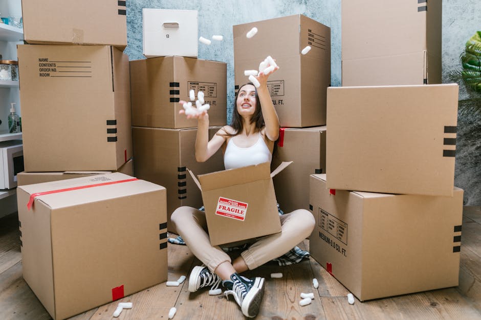 A vintage white delivery van in motion on a roadway, loaded with assorted household furniture and belongings, including wooden chairs, a small table, cardboard boxes, and plastic bags. The furniture i