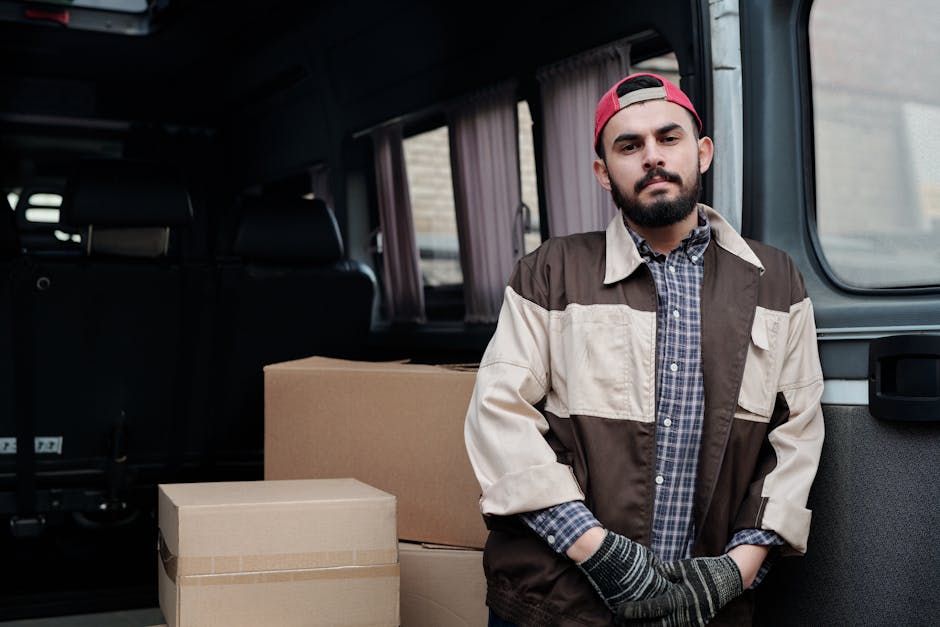 A male mover with a beard and wearing a red cap, a brown and beige jacket over a plaid shirt, stands inside a vehicle with an open rear door. Behind him, several cardboard boxes of various sizes are p